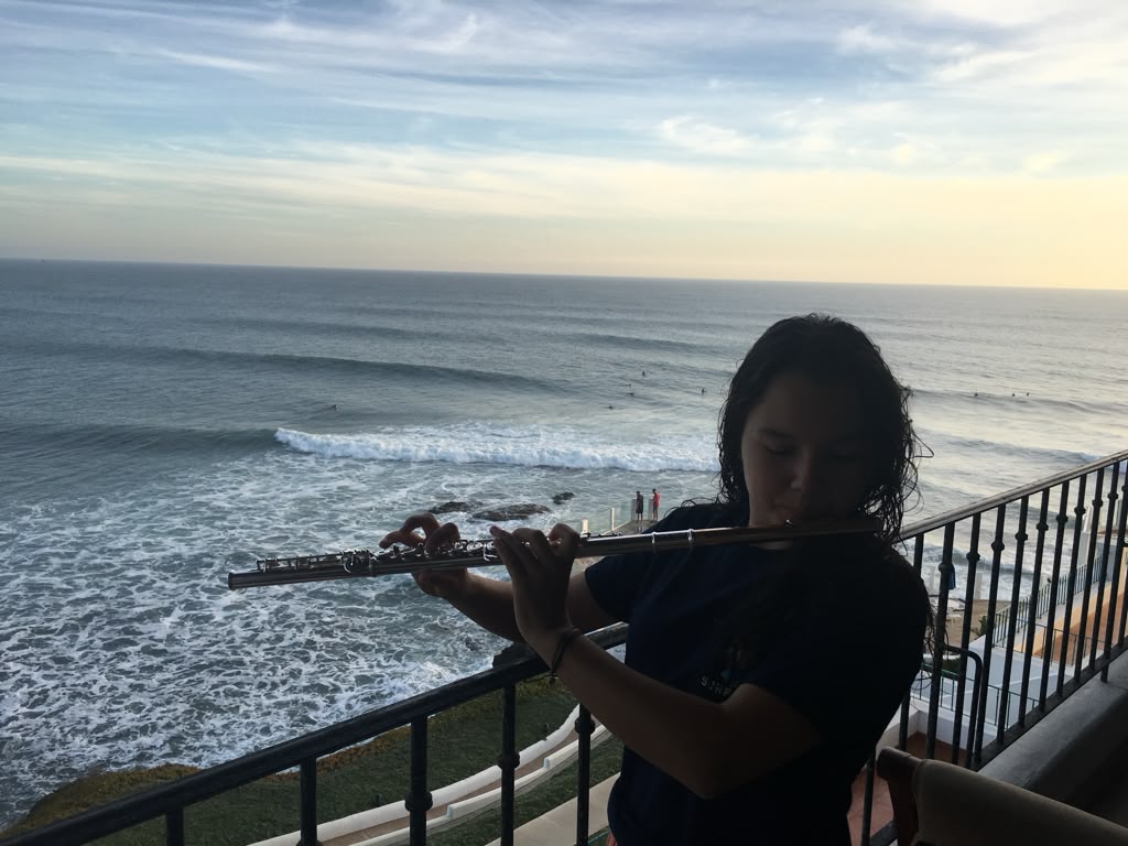 Gabriela Beltrán playing flute on a balcony overlooking the ocean at sunset