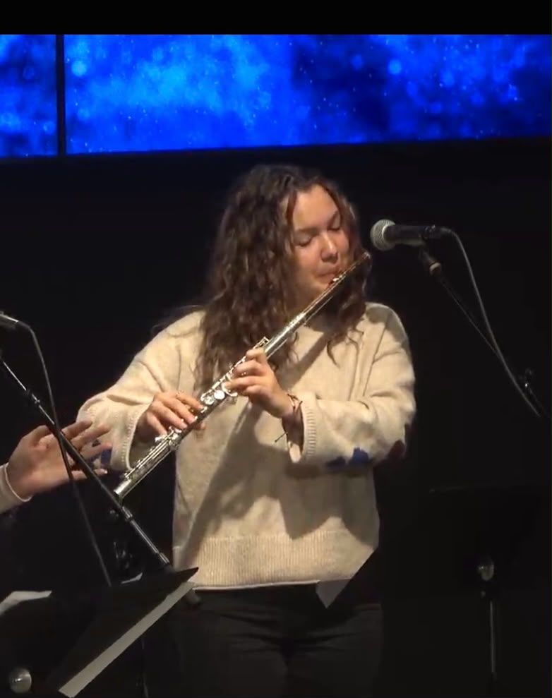 Gabriela Beltrán performing flute on stage with a microphone and blue backdrop