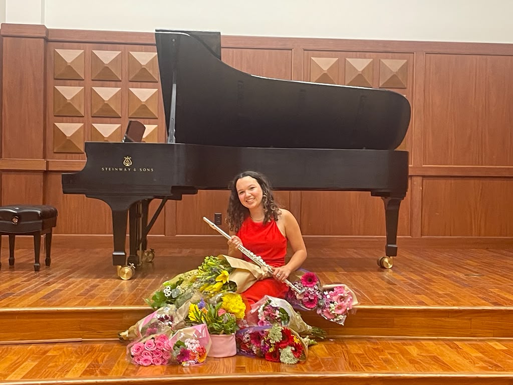 Gabriela Beltrán in a red gown seated with her flute and bouquets after a recital