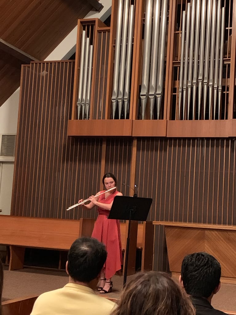 Gabriela Beltrán in a coral dress performing flute before a large pipe organ