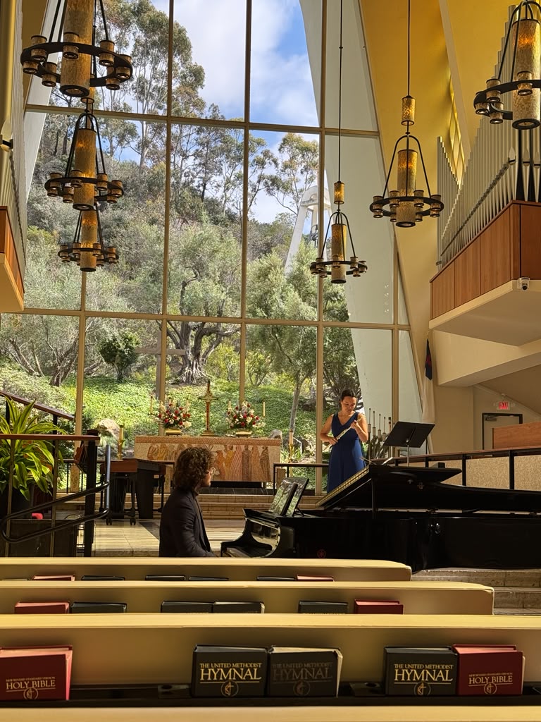 Gabriela Beltrán performing flute with piano in a sunlit chapel with tall windows and a pipe organ