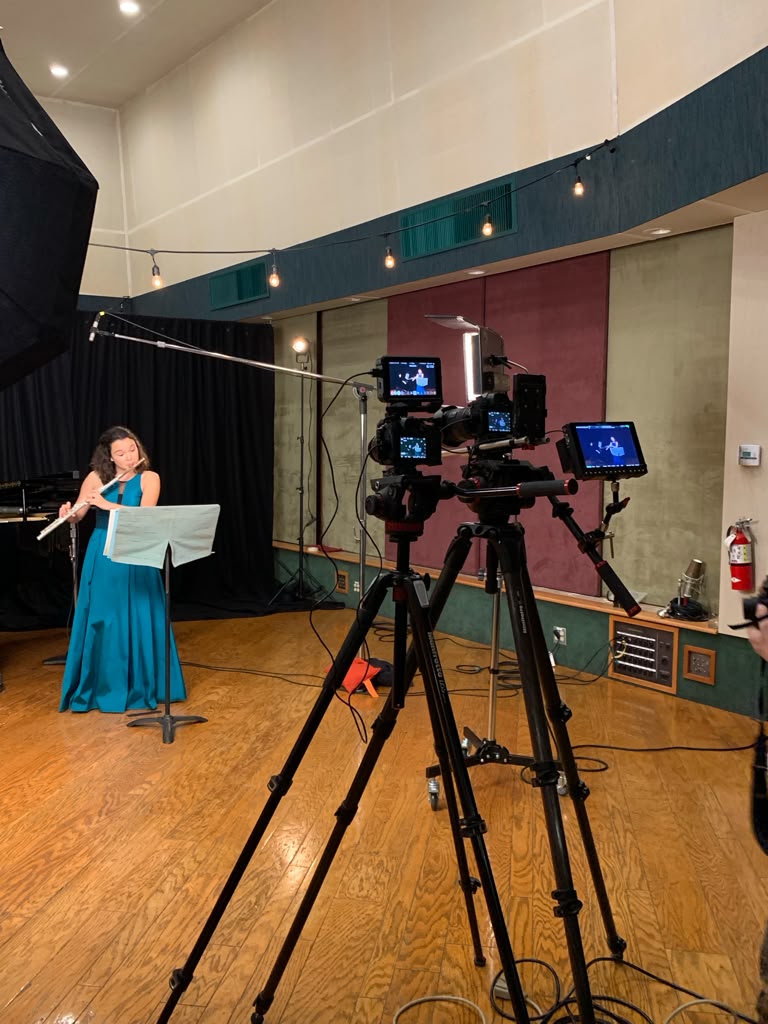 Gabriela Beltrán in a teal gown recording a flute performance on a soundstage with cameras and boom mic