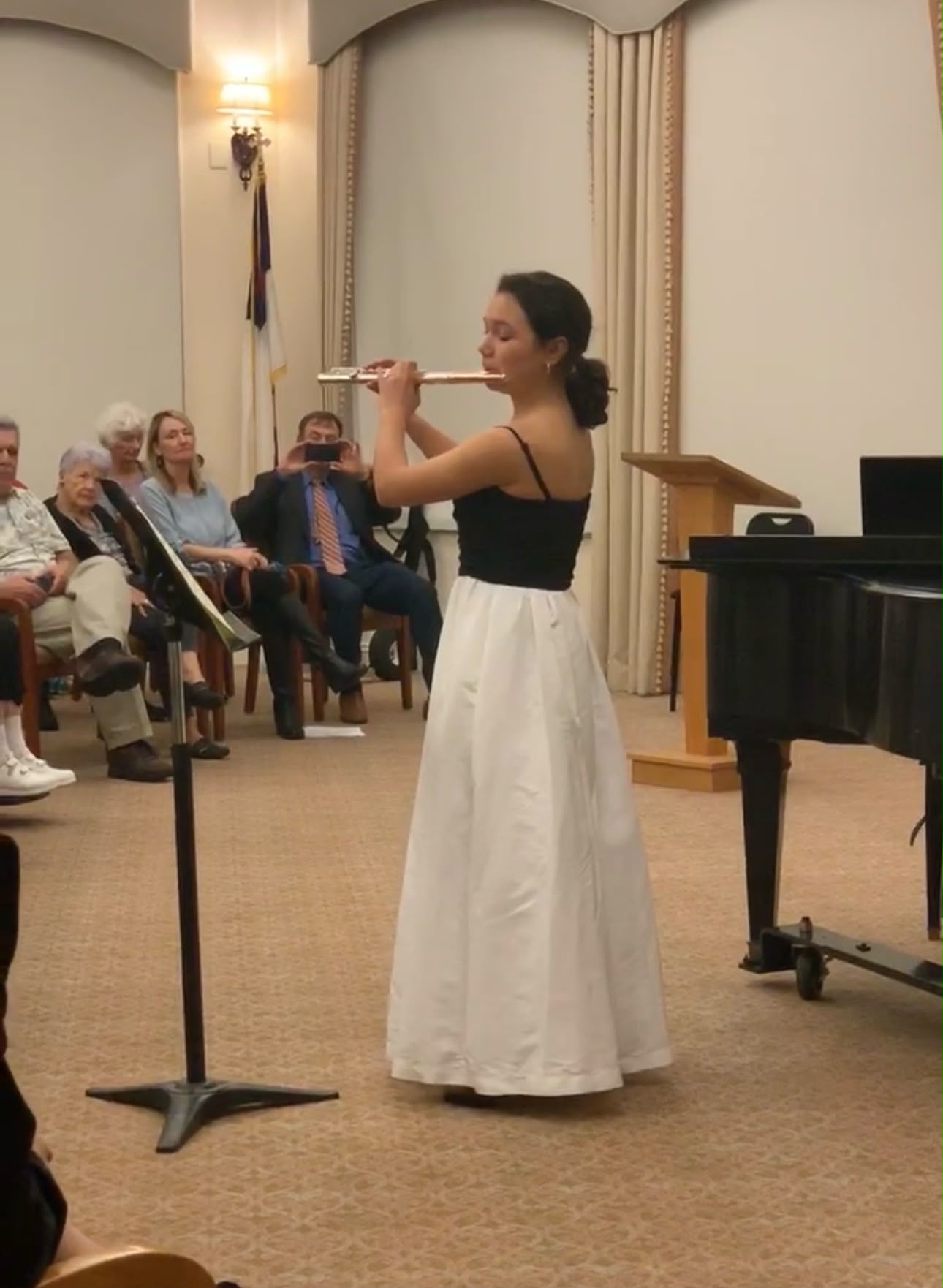 Gabriela Beltrán performing flute in a white gown before a seated audience