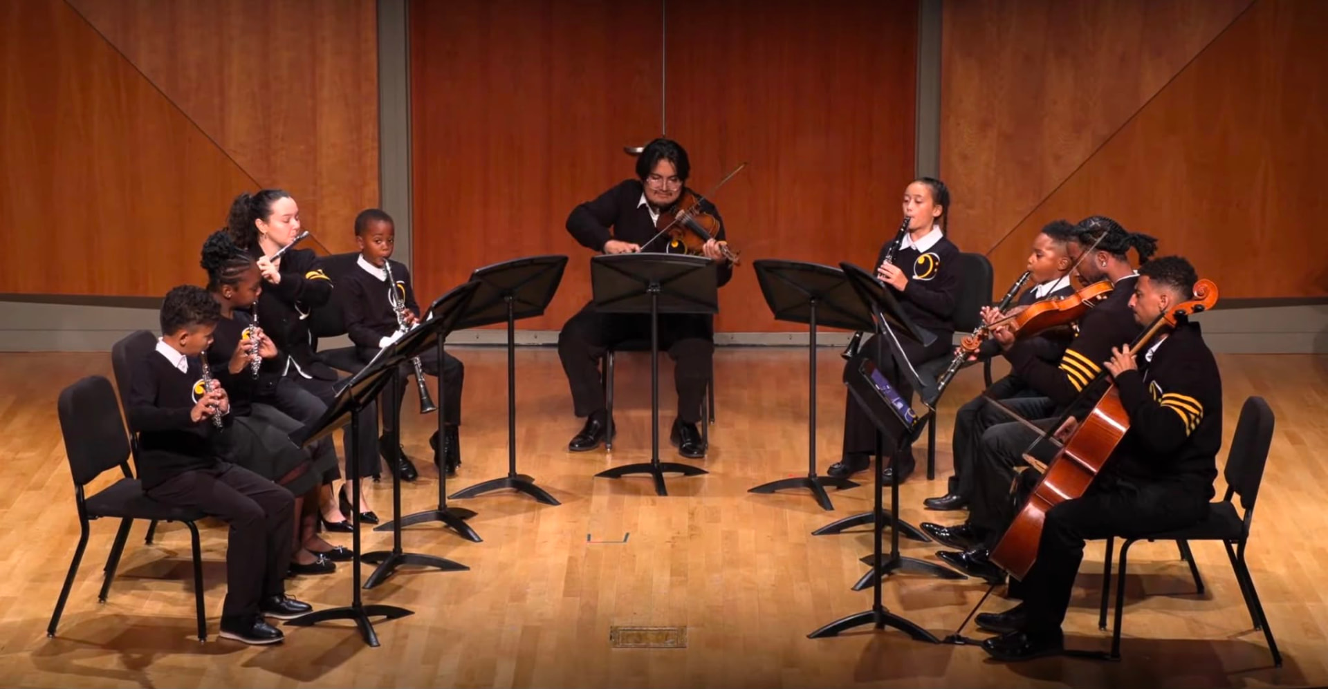 Gabriela Beltrán performing on flute alongside young student musicians in an orchestral ensemble on stage