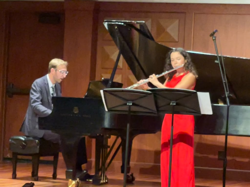 Gabriela Beltrán in a red gown performing flute with piano accompaniment on a recital stage